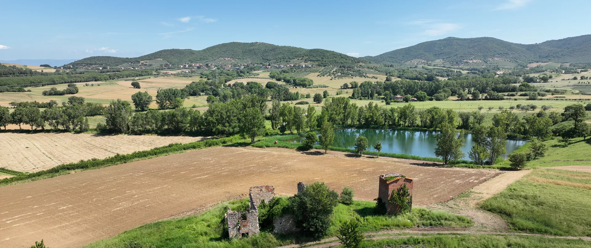 Veduta aerea del paesaggio di Monte Solare: colline boscose, un piccolo lago tra gli alberi, un campo arato e rovine in pietra in primo piano